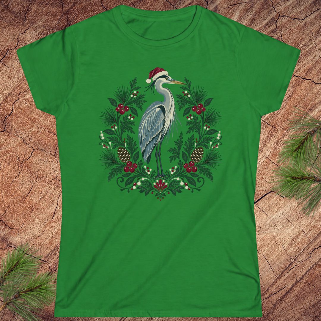 Irish green women's t-shirt with a Great Blue Heron in a Santa hat inside a folk-art wreath of pine, red berries, and pinecones, laid flat on a rustic wood background with pine branches.
