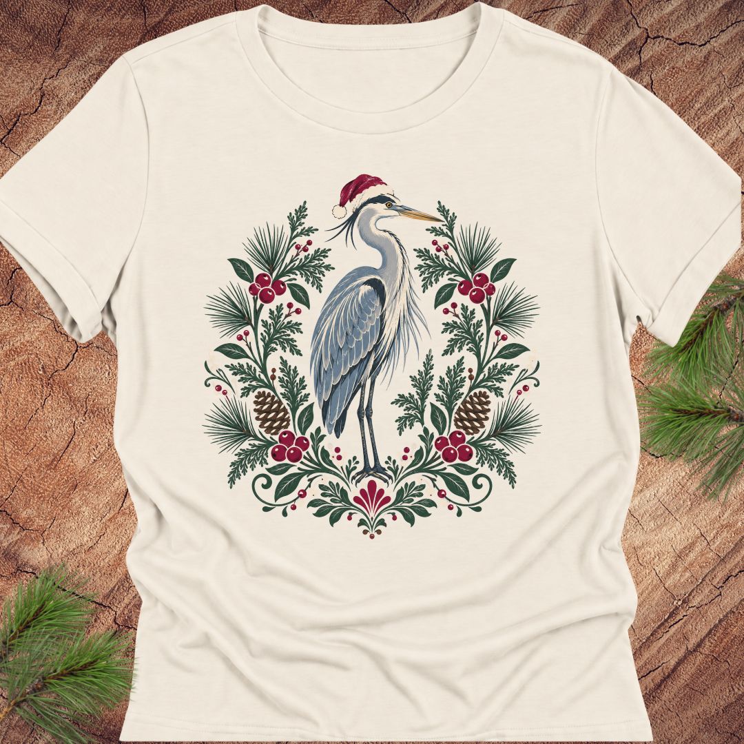 An off-white short-sleeve crewneck t-shirt featuring a festive graphic of a blue heron wearing a red Santa hat, surrounded by a wreath of green pine needles, pinecones, and red berries, laid flat on a rustic wood background with pine branches.