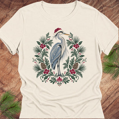 An off-white short-sleeve crewneck t-shirt featuring a festive graphic of a blue heron wearing a red Santa hat, surrounded by a wreath of green pine needles, pinecones, and red berries, laid flat on a rustic wood background with pine branches.