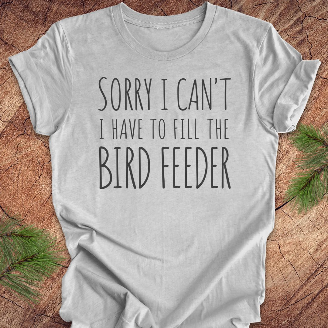A light gray short-sleeve t-shirt with the text 'SORRY I CAN'T I HAVE TO FILL THE BIRD FEEDER' printed in black. The shirt is laid flat on a rustic wood background with green pine branches in the corners.