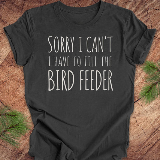A dark charcoal gray t-shirt with white text reading 'SORRY I CAN'T I HAVE TO FILL THE BIRD FEEDER' laid flat on a rustic wood background with pine branches in the corners.