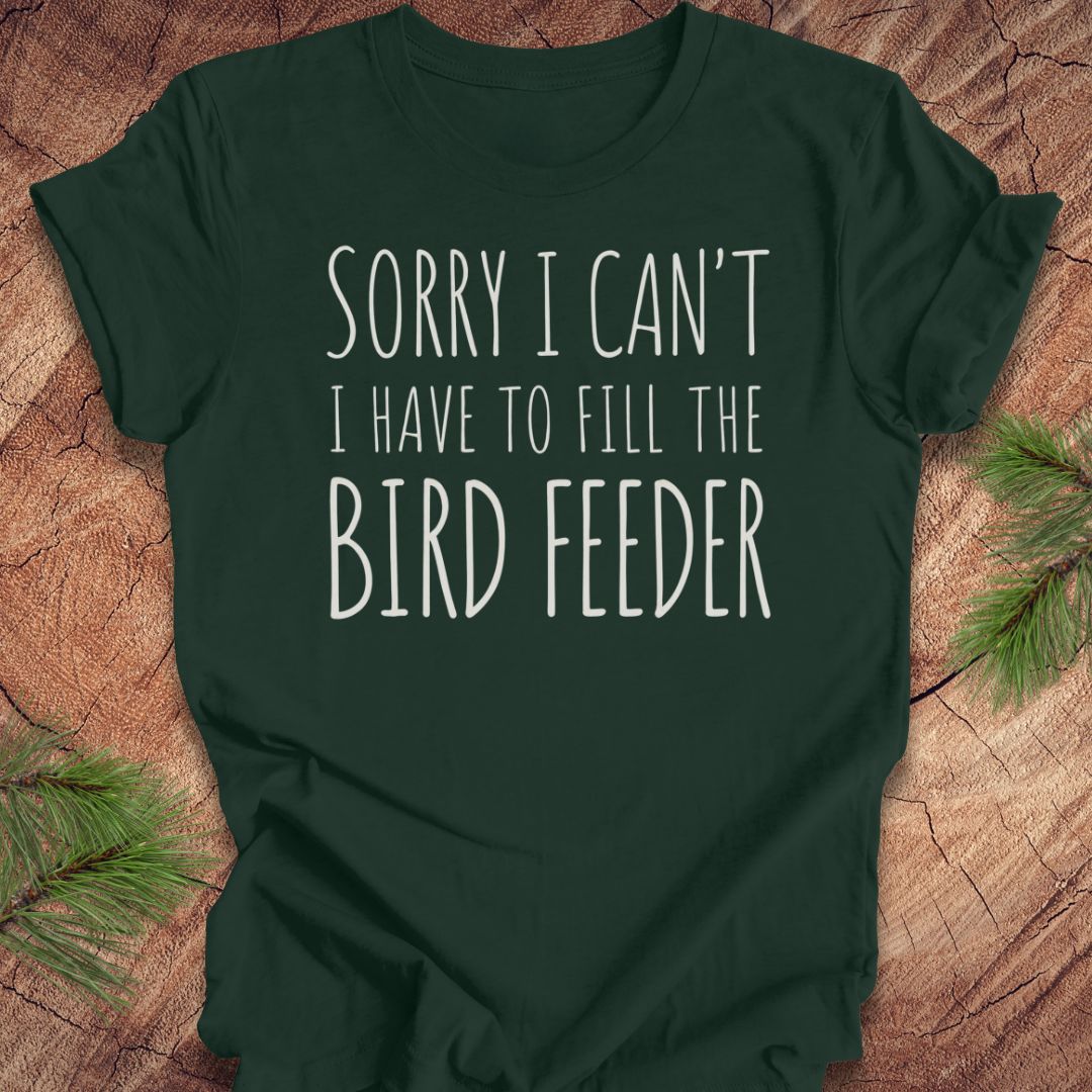 A dark forest green short-sleeve t-shirt with white text reading 'SORRY I CAN'T I HAVE TO FILL THE BIRD FEEDER' printed on the front, laid flat on a rustic wood background with pine branches in the corners.