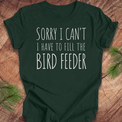 A dark forest green short-sleeve t-shirt with white text reading 'SORRY I CAN'T I HAVE TO FILL THE BIRD FEEDER' printed on the front, laid flat on a rustic wood background with pine branches in the corners.