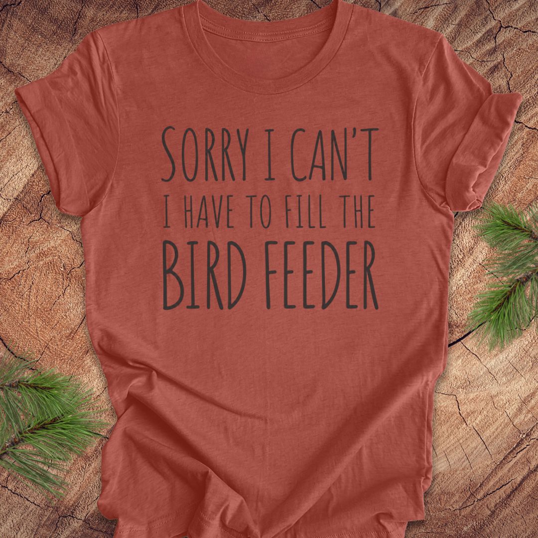 A terracotta-colored short-sleeve t-shirt with the text 'SORRY I CAN'T I HAVE TO FILL THE BIRD FEEDER' printed in dark gray. The shirt is laid flat on a rustic wood slice background with green pine branches in the corners.
