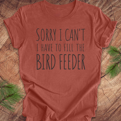 A terracotta-colored short-sleeve t-shirt with the text 'SORRY I CAN'T I HAVE TO FILL THE BIRD FEEDER' printed in dark gray. The shirt is laid flat on a rustic wood slice background with green pine branches in the corners.