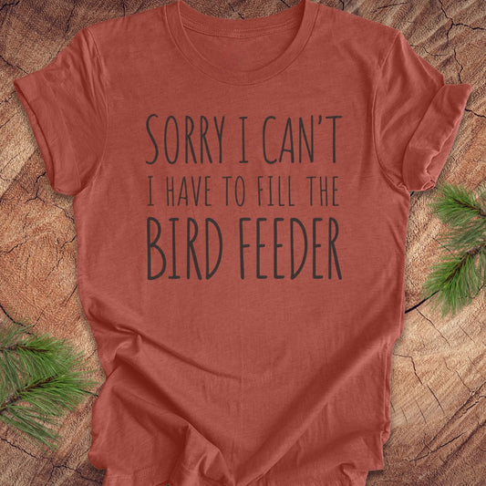 A terracotta-colored short-sleeve t-shirt with the text 'SORRY I CAN'T I HAVE TO FILL THE BIRD FEEDER' printed in dark gray. The shirt is laid flat on a rustic wood slice background with green pine branches in the corners.