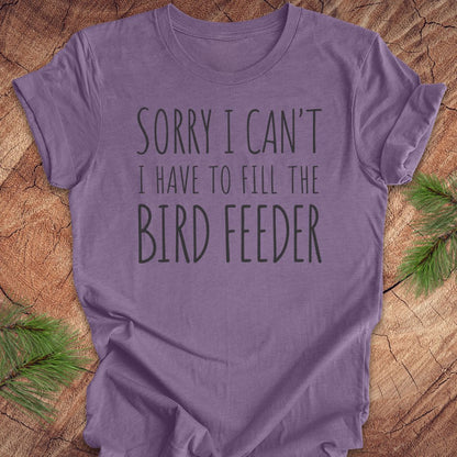A purple short-sleeve t-shirt with the text 'SORRY I CAN'T I HAVE TO FILL THE BIRD FEEDER' printed in dark gray, laid flat on a rustic wood background with pine branches in the corners.