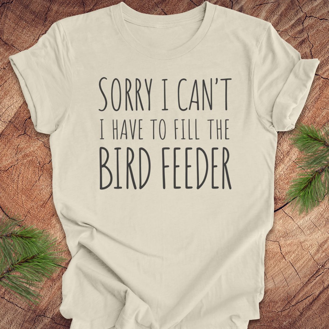 An off-white t-shirt with the text 'SORRY I CAN'T I HAVE TO FILL THE BIRD FEEDER' printed in black, laid flat on a rustic wood background with pine branches in the corners.
