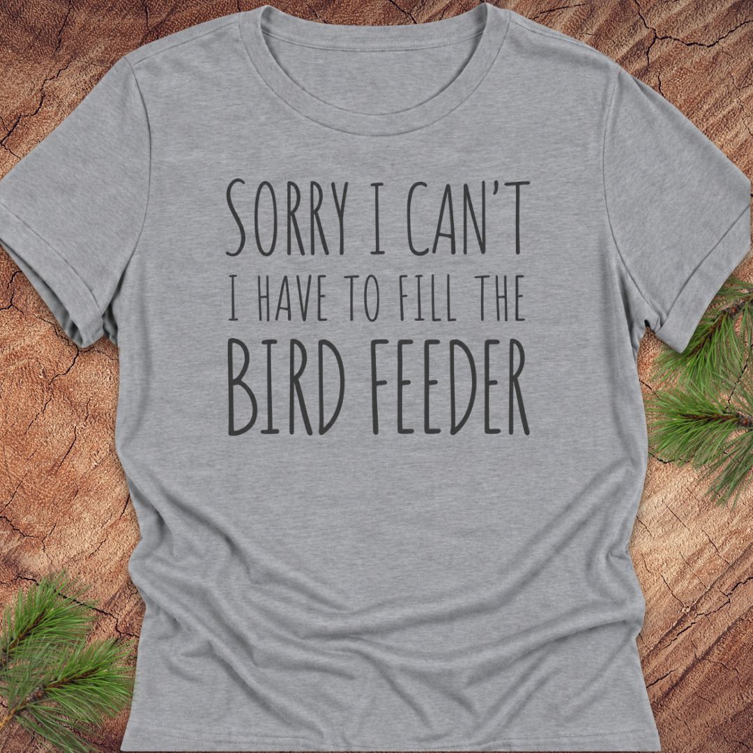 A heather gray short-sleeve t-shirt with the text 'SORRY I CAN'T I HAVE TO FILL THE BIRD FEEDER' printed in black on the front, laid flat on a rustic wood background with pine needles in the corners.