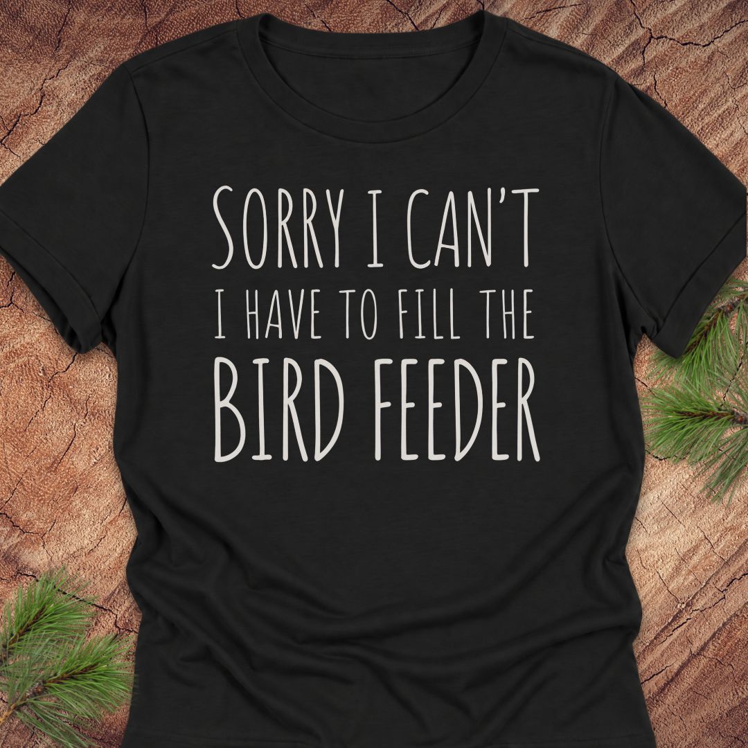 A black short-sleeve t-shirt with white text reading 'SORRY I CAN'T I HAVE TO FILL THE BIRD FEEDER' laid flat on a rustic wood background with pine branches in the corners.