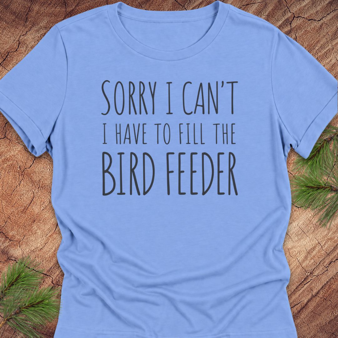 A light blue short-sleeve t-shirt with the text 'SORRY I CAN'T I HAVE TO FILL THE BIRD FEEDER' printed in black on the front, laid flat on a rustic wood background with pine branches in the corners.