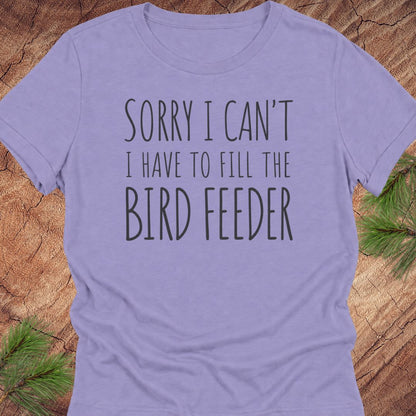 A light purple short-sleeve t-shirt with the text 'SORRY I CAN'T I HAVE TO FILL THE BIRD FEEDER' printed in dark gray. The shirt is laid flat on a rustic wood background with pine needles visible in the corners.