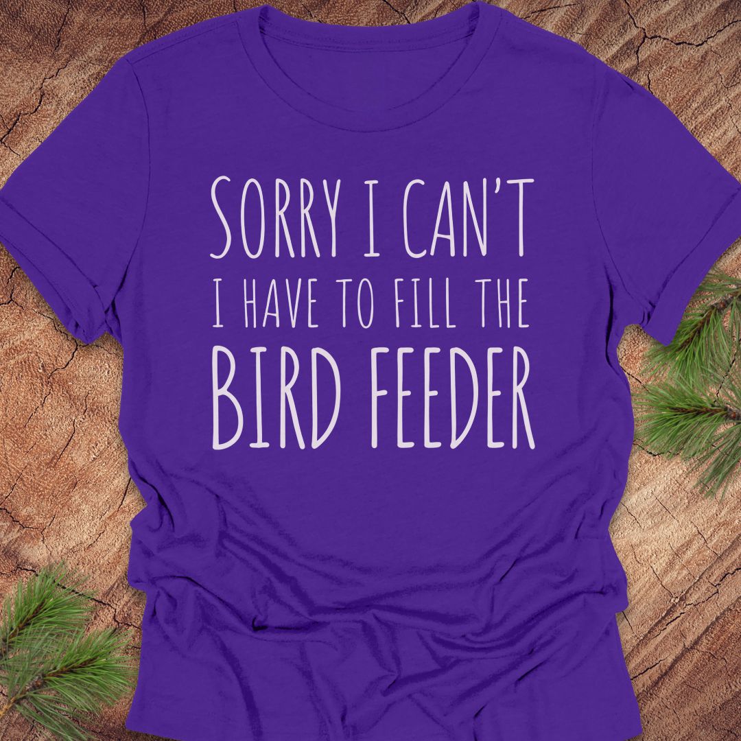 A purple short-sleeve t-shirt with the text 'SORRY I CAN'T I HAVE TO FILL THE BIRD FEEDER' printed in white, laid flat on a rustic wood background with pine branches in the corners.