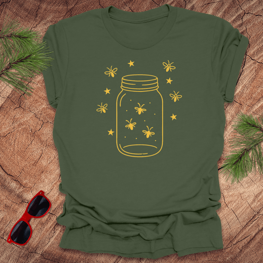 Green t-shirt with a yellow jar and firefly design on a wooden surface with sunglasses and greenery.