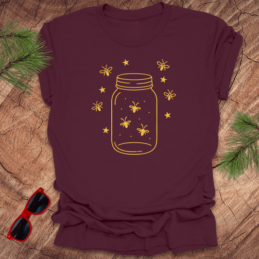 Maroon t-shirt with a yellow jar and firefly design on a wooden surface with sunglasses and greenery.
