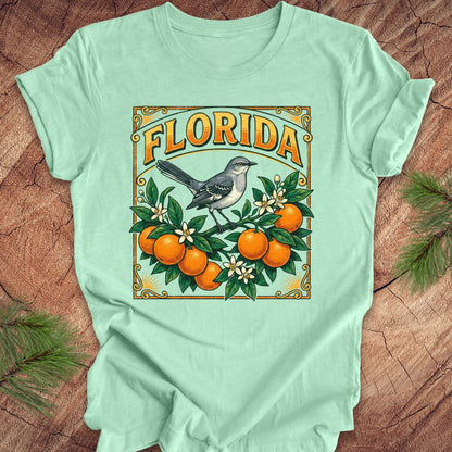 Mint green t-shirt with Florida-themed design featuring oranges and a bird on a wooden surface.