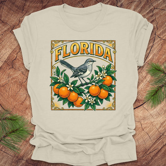 Beige t-shirt with a colorful Florida design with a mockingbird and oranges on a wood background.