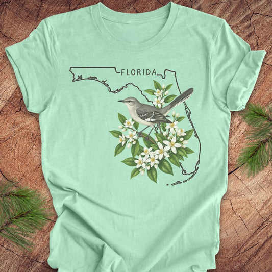 A mint green short-sleeve t-shirt laid flat on a rustic wood surface, featuring a graphic of the Florida state outline with a mockingbird perched among white flowers and green leaves. Pine branches are placed around the shirt as styling props.