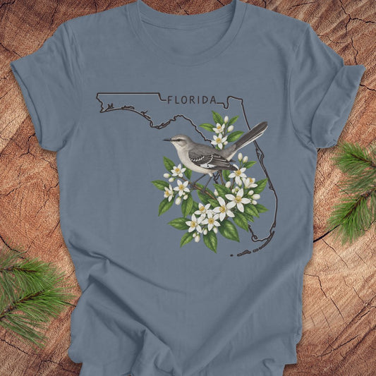A slate blue short-sleeve t-shirt laid flat on a rustic wood background with pine branches in the corners. The shirt features a graphic print of the Florida state outline, a gray-and-white bird perched on green leaves, and small white flowers.