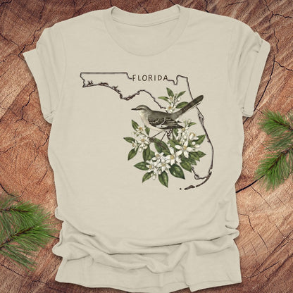 Beige t-shirt with Florida outline, mockingbird, and flowers on a wooden surface
