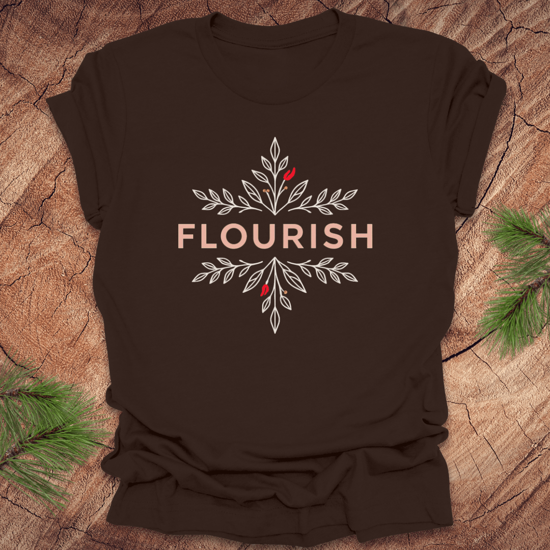 Brown t-shirt with 'FLOURISH' design and flowers on a wooden surface with greenery.