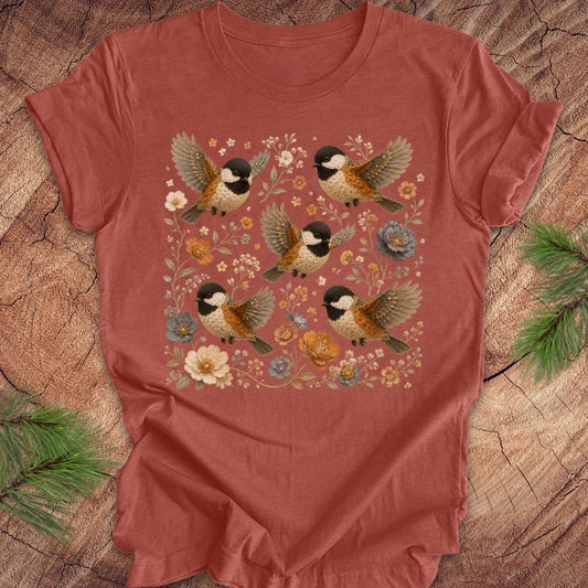 Clay colored t-shirt with chickadees and flowers on a wood background.