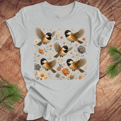 Gray t-shirt with chickadees and flowers on a wood background.