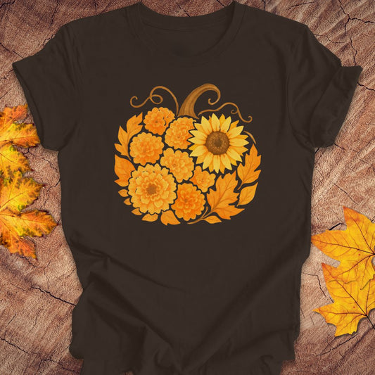 A brown short-sleeve t-shirt laid flat on a rustic wood surface, featuring a large autumn floral pumpkin graphic in orange, yellow, and brown tones. Decorative yellow and orange leaves are placed around the shirt.