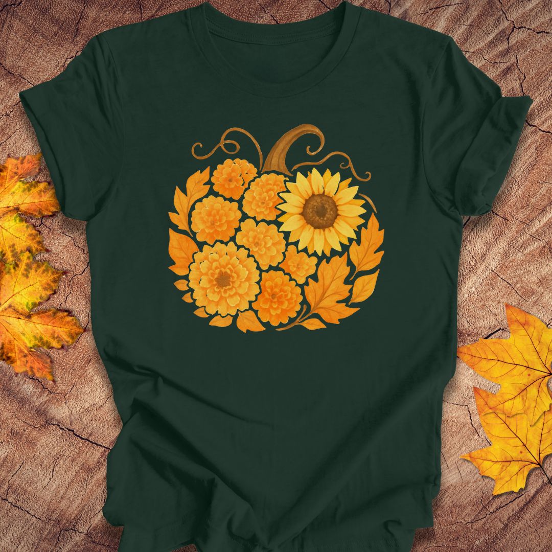 A dark forest green short-sleeve t-shirt with a graphic of a pumpkin made of yellow and orange flowers, laid flat on a rustic wood background with autumn leaves.