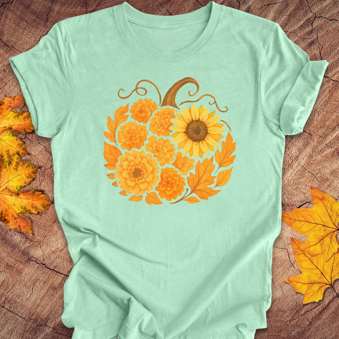 A mint green short-sleeve t-shirt with a graphic of a pumpkin made of orange marigolds and autumn leaves, laid flat on a rustic wood background with scattered yellow leaves.