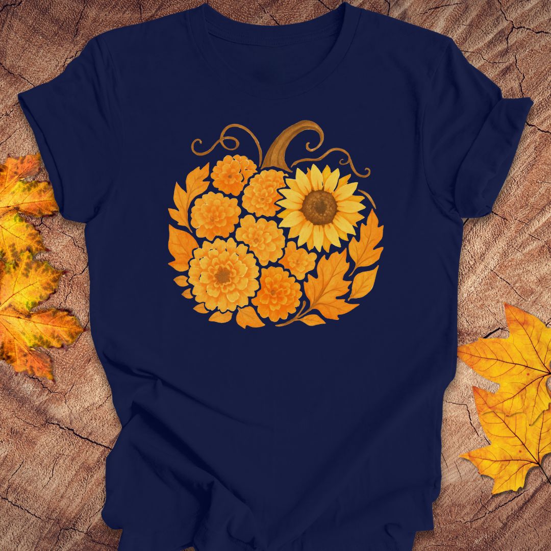 A navy blue short-sleeve t-shirt with a graphic of a pumpkin made of yellow and orange flowers, laid flat on a rustic wood background with autumn leaves.