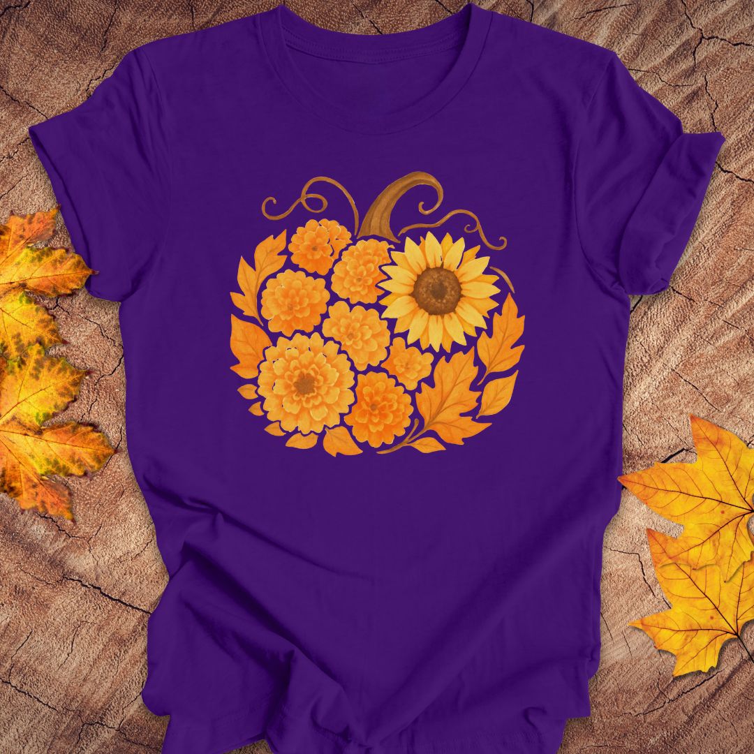 A purple short-sleeve t-shirt laid flat on a rustic wood surface, decorated with a large graphic of orange marigolds and a yellow sunflower arranged in a pumpkin shape. Yellow and orange autumn leaves are scattered around the edges of the frame.
