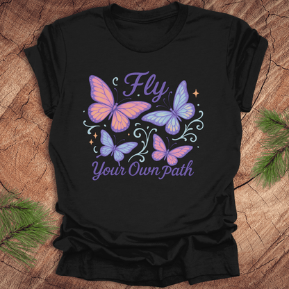 Black t-shirt with butterfly design and 'Fly Your Own Path' text on a wooden surface.