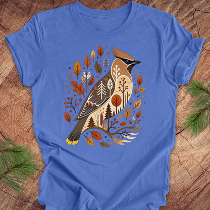 A blue short-sleeve crewneck t-shirt laid flat on a rustic wood surface, featuring a large graphic of a bird surrounded by autumn leaves and small trees.