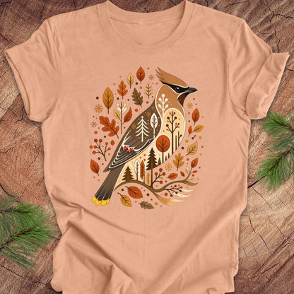 A peach-colored short-sleeve t-shirt with a detailed graphic of a cedar waxbird surrounded by autumn leaves and pine trees, laid flat on a rustic wood background with pine branches.