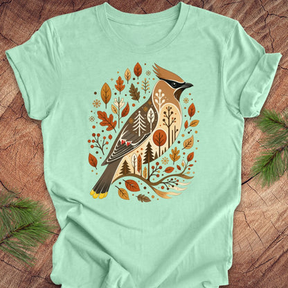 A mint green short-sleeve t-shirt laid flat on a rustic wood background, featuring a detailed graphic of a cedar waxbird surrounded by autumn leaves and pine trees.