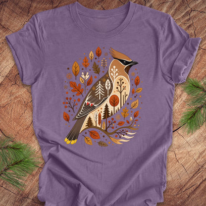 A purple short-sleeve crewneck t-shirt featuring a detailed graphic of a cedar waxbird surrounded by autumn leaves and pine trees, laid flat on a rustic wood background with pine needles in the corners.