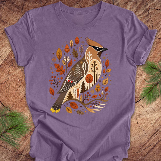 A purple short-sleeve crewneck t-shirt featuring a detailed graphic of a cedar waxbird surrounded by autumn leaves and pine trees, laid flat on a rustic wood background with pine needles in the corners.