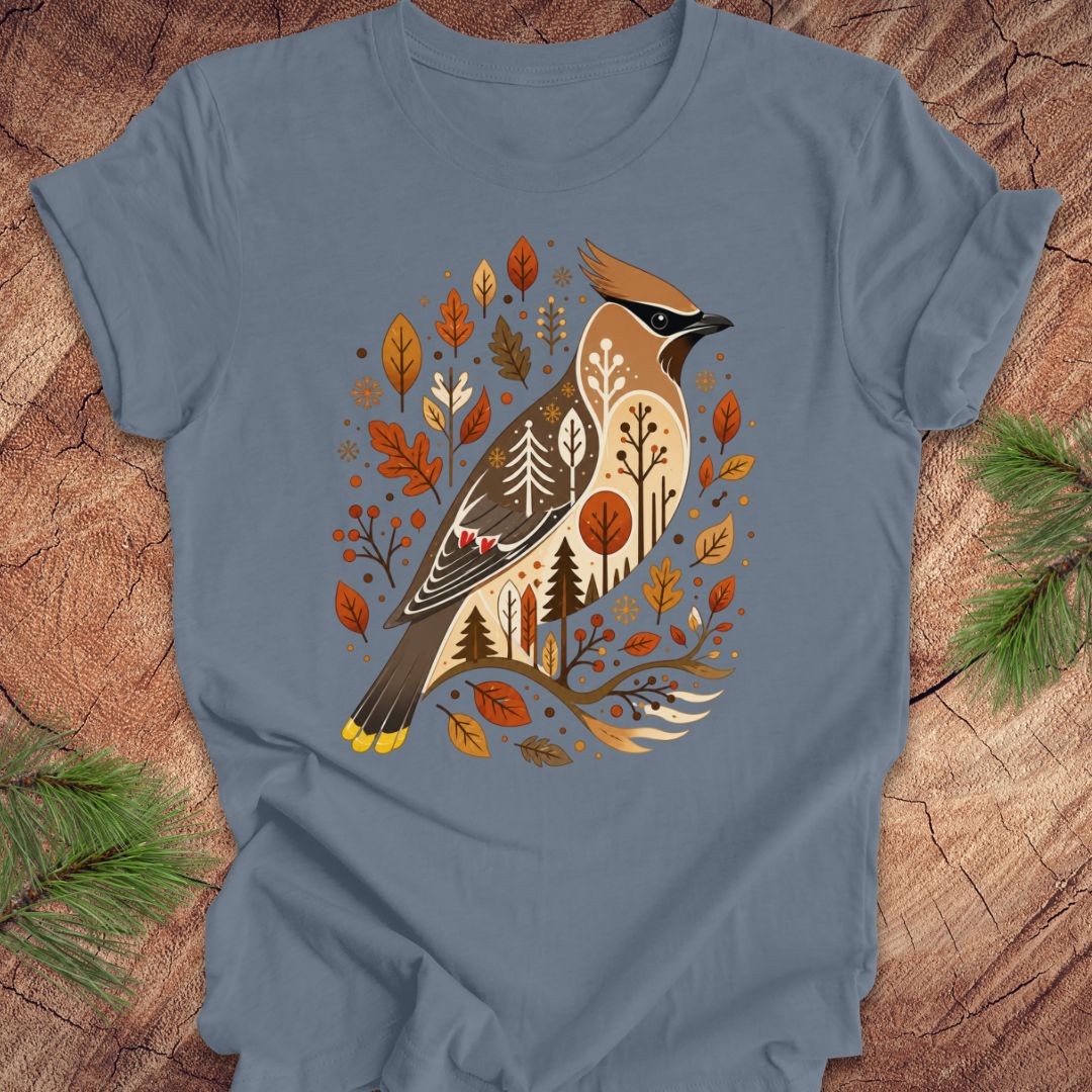 A slate blue crewneck t-shirt featuring a detailed graphic of a cedar waxwing bird surrounded by autumn leaves and pine trees, laid flat on a rustic wood background with pine needles in the corners.