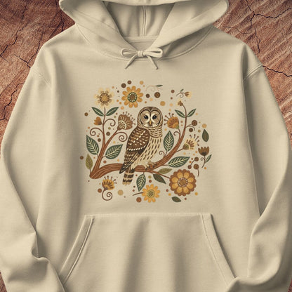 An off-white hooded sweatshirt with a detailed graphic of an owl perched on a branch surrounded by autumn leaves, sunflowers, and dots, displayed against a textured brown wood background.
