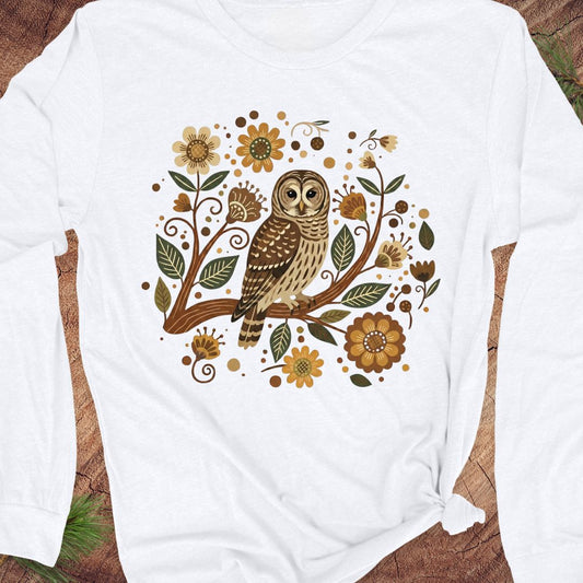 A white long-sleeve t-shirt with a graphic print of a brown owl perched on a branch surrounded by autumn flowers and leaves, laid flat on a rustic wood background.