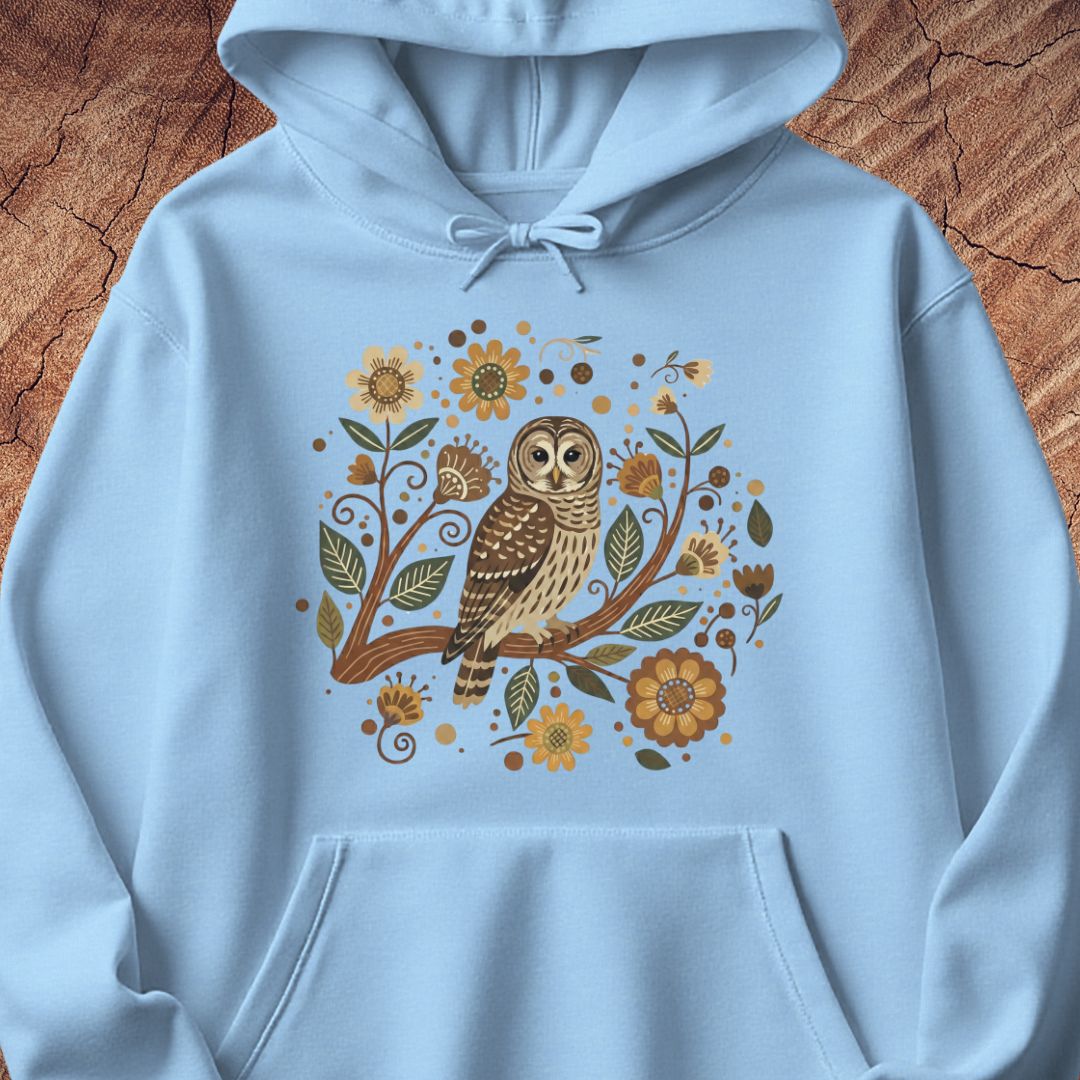 A light blue pullover hoodie laid flat on a textured brown wood background, featuring a central graphic of a brown owl perched among yellow flowers and green leaves.