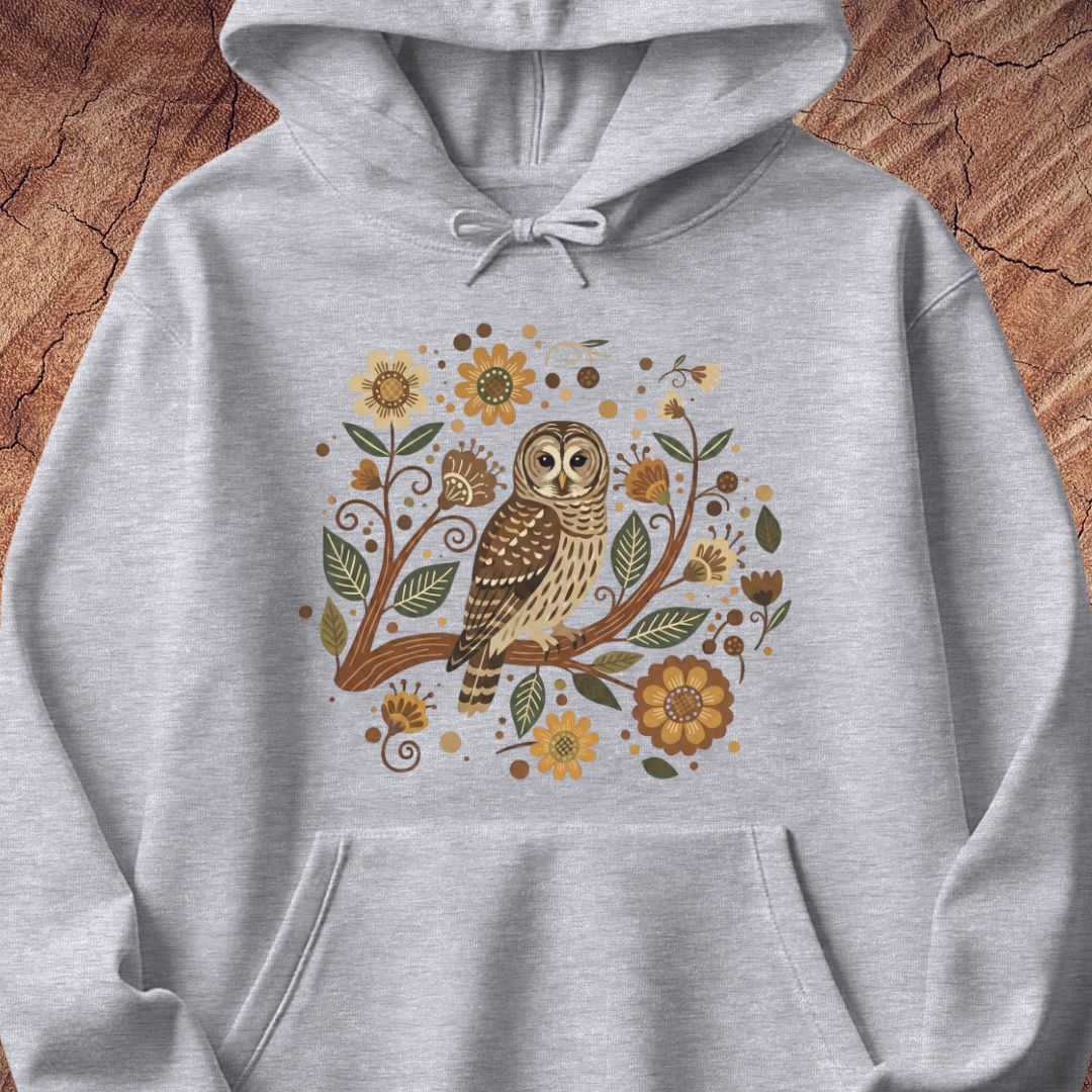 A light heather gray pullover hoodie featuring a central graphic of a brown and beige owl perched on a branch surrounded by yellow flowers and green leaves, displayed against a rustic wood background.