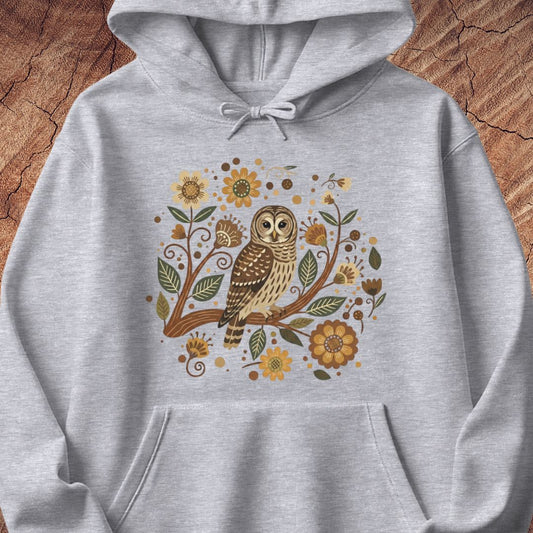 A light heather gray pullover hoodie featuring a central graphic of a brown and beige owl perched on a branch surrounded by yellow flowers and green leaves, displayed against a rustic wood background.