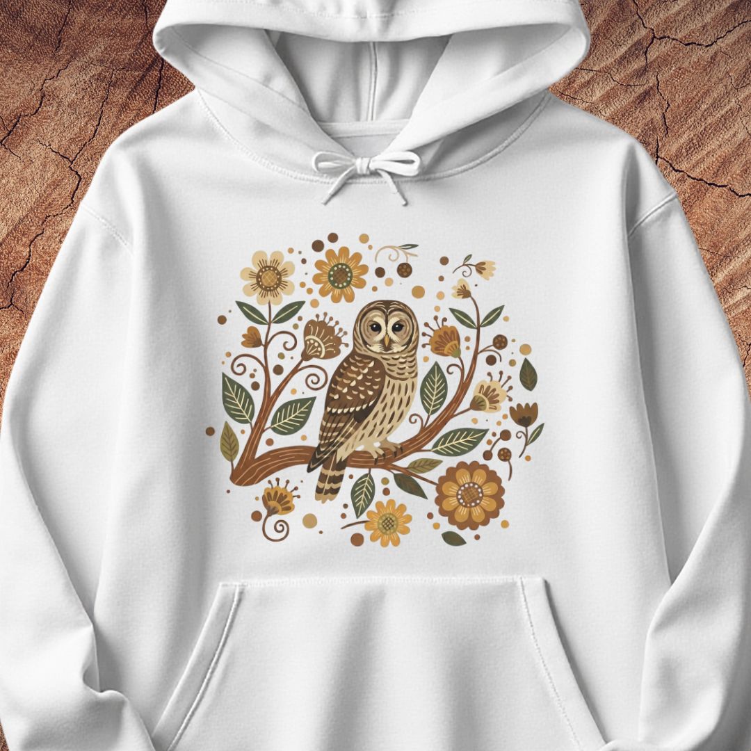 A white pullover hoodie featuring a detailed graphic of a brown and beige owl perched on a branch surrounded by autumn leaves and flowers, displayed on a textured brown wood background.