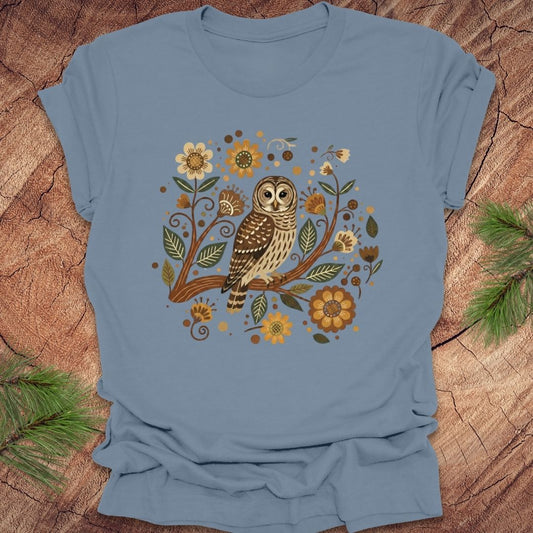 Steel blue t-shirt with a barred owl on a branch surrounded by folk art style flowers, on a wooden background.