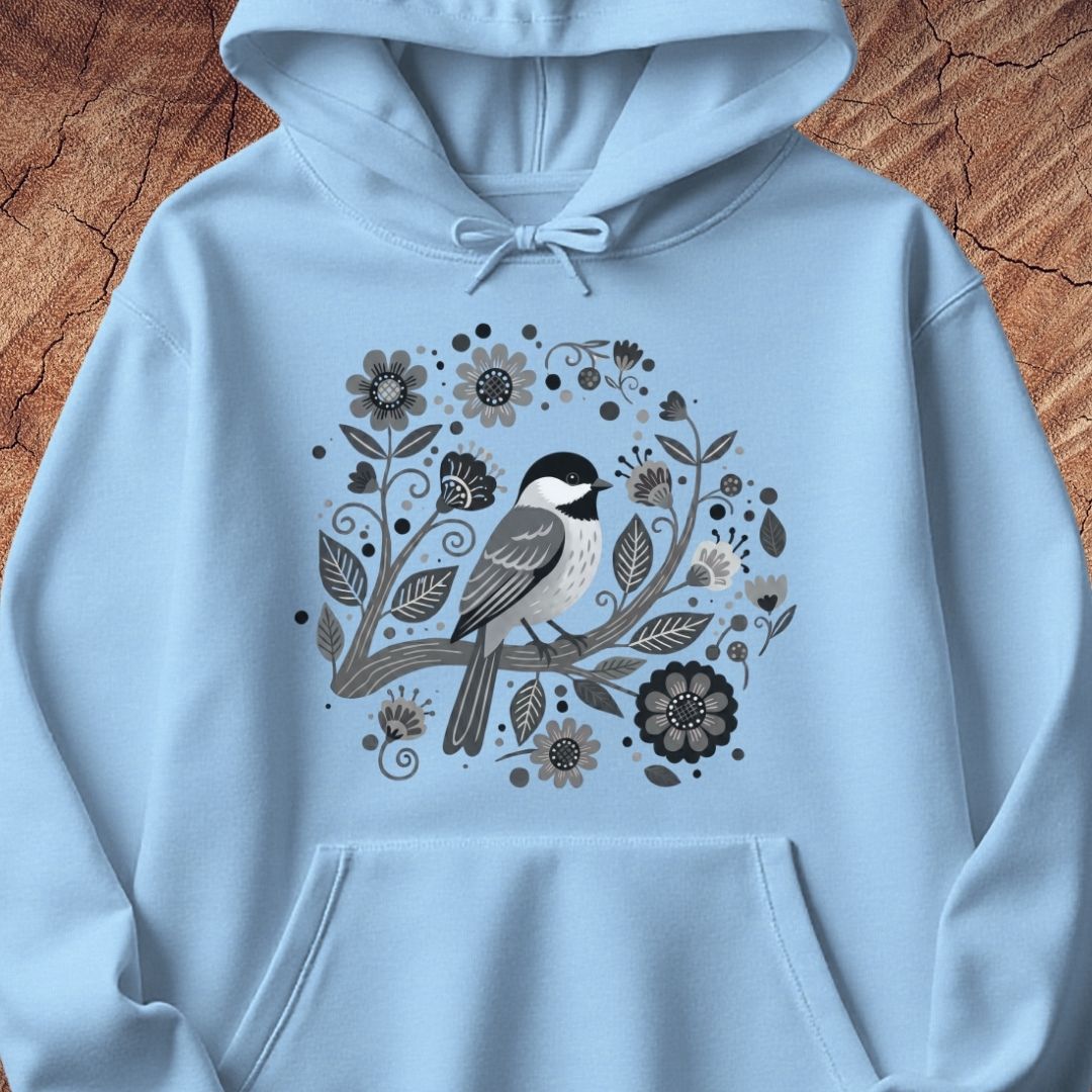 A light blue pullover hoodie featuring a central graphic of a black-capped chickadee perched among stylized gray and black flowers and leaves, displayed flat against a rustic wood background.