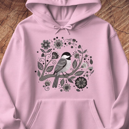 A light pink pullover hoodie featuring a central graphic of a black-capped chickadee surrounded by black, gray, and white floral and leafy illustrations, displayed flat against a rustic wood background.