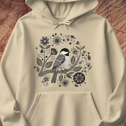 A cream-colored pullover hoodie featuring a central graphic of a black-capped chickadee bird surrounded by stylized black and gray flowers and leaves, displayed on a textured brown wood background.