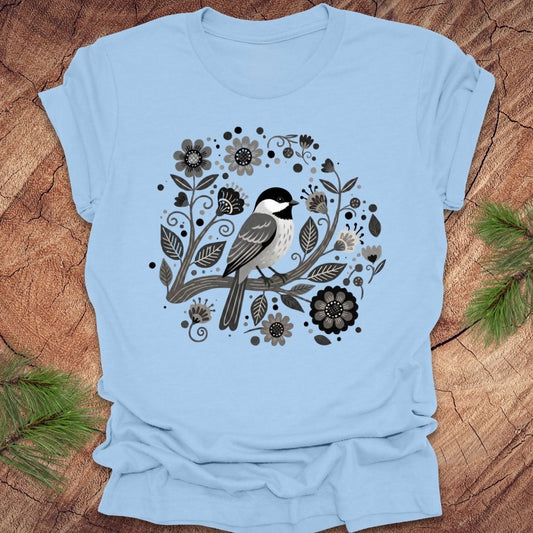 Baby blue t-shirt of a chickadee on a branch surrounded by folk art flowers, all in black and white, on a wooden background.